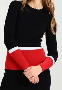 Schwarzes, geripptes Langarmoberteil mit einem rot-weißen gestreiften Bund und Ärmelbündchen, das ein figurnahes Design und ein strukturiertes Material hervorhebt.