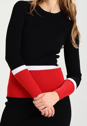 Haut noir à manches longues à côtes avec une taille et des poignets rayés rouges et blancs, mettant en avant une coupe ajustée et un matériau texturé.