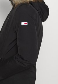 Svart utomhusjacka med en huva fodrad med päls. Tyget verkar slätt och har en logotyp på ärmen. Dragkedjedetaljer är synliga.