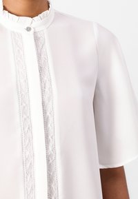 Blusa bianca con colletto alto arricciato, dettaglio centrale con bottoni e raffinato pizzo lungo la parte anteriore e le maniche. Tessuto setoso.