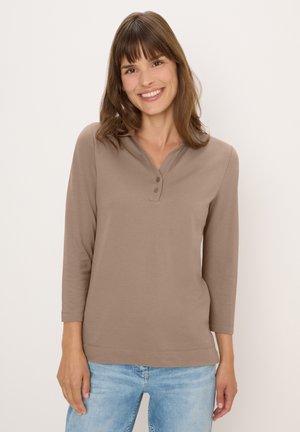 Femme souriante aux cheveux bruns, portant un haut taupe à manches longues avec des boutons et un jean bleu clair sur fond uni.