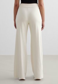 Wijde broek in crèmekleurige stof, met een gladde textuur, hoge tailleband en strakke lijnen. Schoenen zijn zichtbaar aan de zoom.