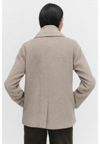 Beige wool-blend coat with a lapel collar, straight cut, and single rear vent, paired with dark textured pants.
