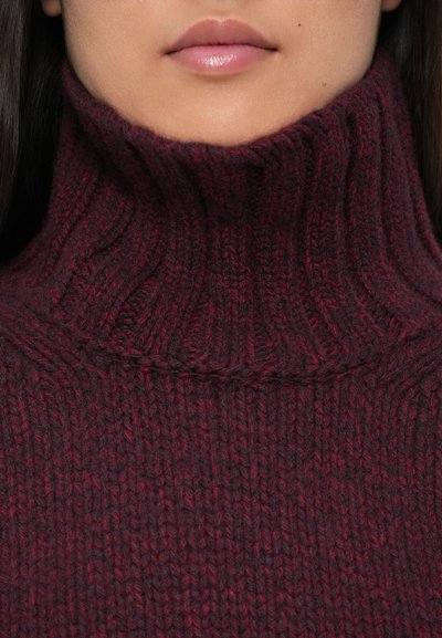 Gros plan sur le bas du visage d'une personne portant un pull à col roulé épais, côtelé, de couleur rouge foncé avec de subtiles variations de teinte.