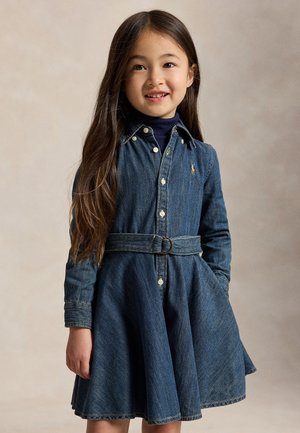 Jeune fille aux longs cheveux foncés portant une robe en denim bleue avec une ceinture et un col roulé sombre en dessous, souriante et debout les mains dans les poches.