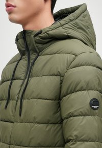 Olívazöld párnázott dzseki kapucnival, cipzárral és fekete húzózsinórral. A bal ujján figyelemre méltó logófolt található. Texturált steppelt dizájn.