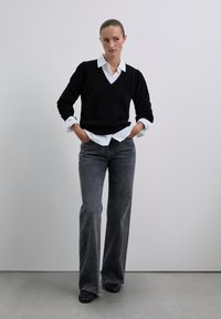 Frau steht mit den Händen in den Taschen, trägt einen schwarzen V-Ausschnitt-Pullover über einem weißen Hemd und weiten, verblassten schwarzen Jeans vor einem einfarbigen Hintergrund.