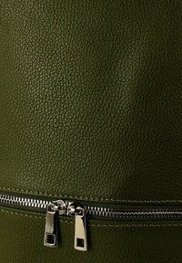 Close-up van donkergroen gestructureerd leer met een horizontale zilveren dubbele ritssluiting die gedeeltelijk aan de onderkant gesloten is.