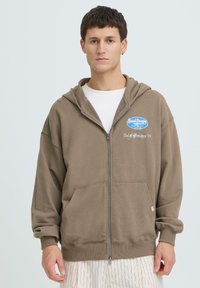 Sweat à capuche marron avec fermeture éclair et poche avant, arborant un logo bleu et un texte blanc. Le matériau semble doux avec une coupe décontractée et des poignets côtelés.