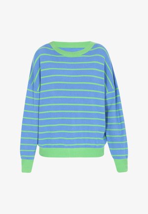 Młoda kobieta patrzy w dół, ubrana w niebiesko-zielony pasiasty sweter, kremowe szerokie spodnie i różowe trampki na białym tle.