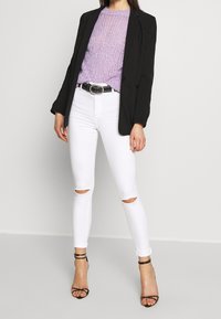 Blazer negro sobre un suéter de punto lavanda. Jeans skinny blancos con rotos y tacones negros de tiras completan el atuendo, creando un look en capas.