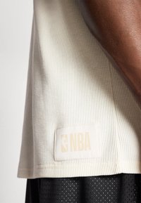 Lys beige teksturert stoffskjorte med et rektangulært NBA-logo i lys beige, båret over en svart mesh-mønstret underdel.