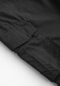 Gros plan sur un tissu noir avec des coutures détaillées et un rabat rectangulaire cousu, probablement une partie d'un vêtement ou d'un sac.