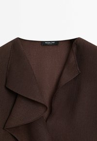 Tissu texturé marron foncé avec un large col volanté asymétrique et une étiquette "Massimo Dutti" visible au niveau de l'encolure.