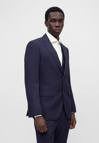 Homme portant un costume formel bleu marine foncé avec une chemise blanche, debout devant un fond clair uni.