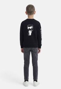 Αγόρι που στέκεται γυρισμένο με την πλάτη, φοράει μαύρο φούτερ με λευκή καρικατούρα και κείμενο στο μανίκι, σκούρα γκρι τζιν και λευκά αθλητικά παπούτσια.