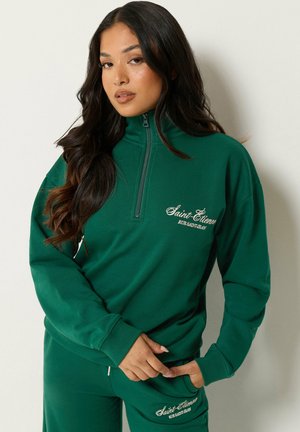 Kvinde med langt mørkt hår iført en grøn half-zip sweatshirt og matchende bukser med hvid Saint-Étienne tekstbroderi.