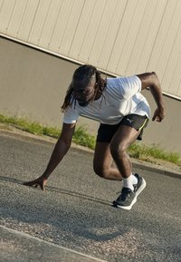 Athlet in einem eng anliegenden weißen Shirt und schwarzen Shorts, hockend auf einer Straße. Trägt schwarze Laufschuhe mit weißen Akzenten und zeigt eine dynamische Haltung.