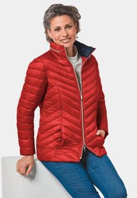 Veste matelassée rouge à motif en chevrons, avec fermeture éclair à l'avant et col haut, associée à un haut gris clair et un jean bleu. Texture lisse.
