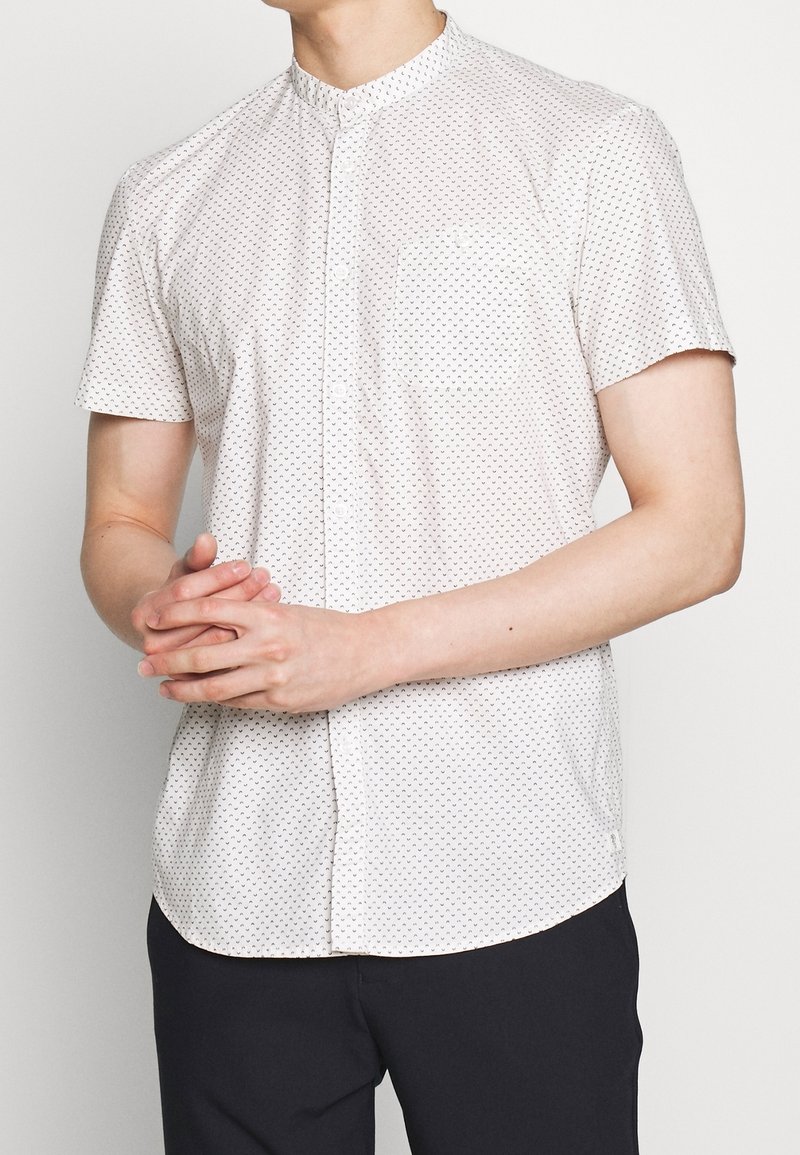 Homme portant une chemise blanche à manches courtes avec un petit motif noir et un pantalon noir, debout les mains jointes.