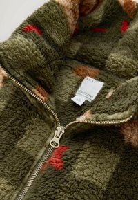 Close-up of partially zipped green fleece jacket with multicolor spots and visible white clothing label inside collar.