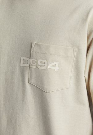 Cremfarbendes Langarmshirt mit einer strukturierten Tasche. Auf der oberen linken Seite befindet sich ein erhabenes weißes Grafikdesign mit der Aufschrift "DC94".