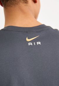 Dunkelgraues Baumwoll-T-Shirt mit einem goldenen Nike-Swoosh-Logo und dem Wort "AIR" in Weiß, das auf dem oberen Rücken platziert ist.