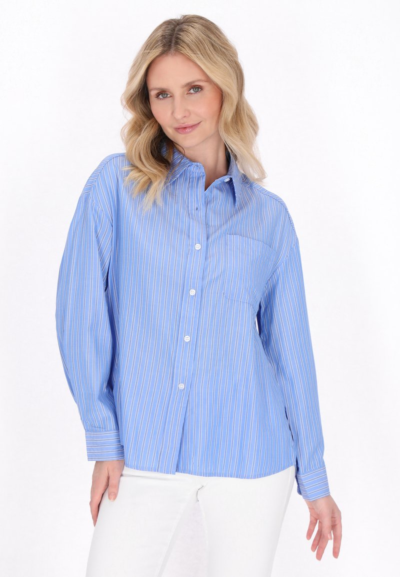 Camicia azzurra chiara con bottoni, caratterizzata da righe verticali bianche, colletto e maniche lunghe con polsini a bottone. Il tessuto appare liscio e leggero.