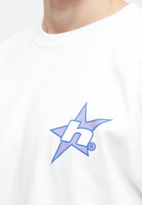 Λευκό βαμβακερό T-shirt με μπλε και μωβ λογότυπο αστέρα που περιλαμβάνει ένα στιλιζαρισμένο "h" στην αριστερή περιοχή του στήθους. Στρογγυλή λαιμόκοψη και κοντά μανίκια.