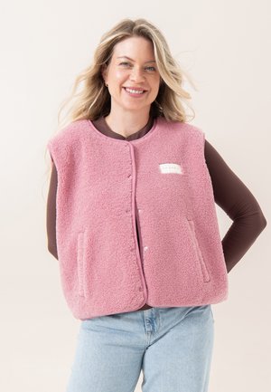 Lächelnde Frau mit langen blonden Haaren, die eine ärmellose pinke Fleeceweste über einem braunen Langarmshirt und hellblauen Jeans trägt.