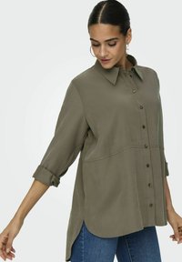 Blusa verde oliva a maniche lunghe con bottoni frontali, colletto strutturato e orlo arrotondato; rifinita con maniche arrotolate e una texture sottile.