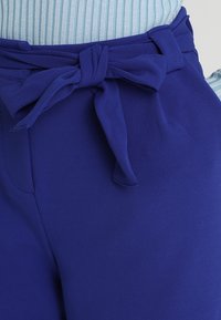 Un pantalon en tissu bleu avec une ceinture à nouer à la taille. Le matériau semble lisse et doux, avec un design à jambes larges.