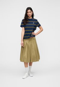 Femme aux longs cheveux foncés portant un t-shirt bleu marine à rayures jaunes, une jupe midi plissée kaki, des chaussettes blanches et des baskets blanches.