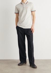 Polo gris clair avec des accents rayés rouges et bleu marine et trois boutons, associé à un pantalon noir et des baskets foncées sur un fond neutre.