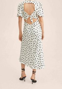 Robe blanche à pois noirs avec de courtes manches bouffantes, dos nu et détail à nouer. Elle présente une jupe fluide et des talons noirs à brides au niveau de la cheville.
