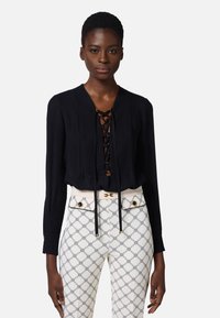 Blusa negra con un frente con cordones, mangas largas y un sutil detalle texturizado, combinada con pantalones crema que presentan un patrón geométrico y acentos dorados.