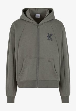 Sweat à capuche gris zippé avec poches avant, poignets et ourlet côtelés, et un logo "K" stylisé noir sur la poitrine gauche.