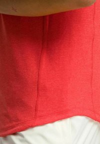 Camiseta de algodón roja con una textura suave, escote redondo y dobladillo curvado. Detalles de costura visibles a lo largo de los lados y los bordes inferiores.