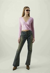 Top cropped rosa de manga longa com design em cruz, combinado com jeans azuis boca de sino. A modelo usa óculos de sol escuros e sapatos de salto agudo castanhos.