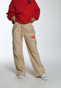 Beige cargobroek van stof met een elastische tailleband, zijzakken en rode grafische accenten. Draagt een rode oversized sweater en sneakers.