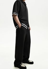 Musta poolopaita, jossa on valkoiset raidalliset hihansuut, yhdistettynä löysiin mustiin housuihin ja kaksivärisiin kenkiin, joissa on teksturoitu päällinen.