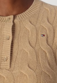 Pull en tricot beige avec un col rond, des bords côtes, trois boutons sur le plastron et un petit logo rouge, blanc et bleu.