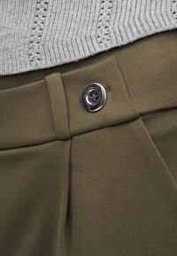 Κοντινή λήψη της μέσης σε παντελόνι ελιάς με ασημί κουμπί τεσσάρων τρυπών και μπεζ τελείωμα πλεκτού πουλόβερ από πάνω.