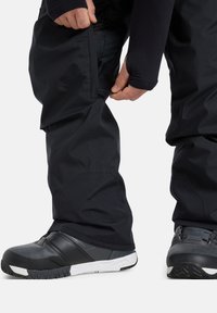 Persoon die de rits van zwarte waterdichte broek aanpast, met zwart-grijze sneeuwlaarzen met witte zolen op een witte achtergrond.
