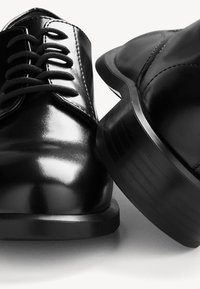 Chaussures de soirée en cuir noir, avec une finition brillante, dotées de lacets, d'un bout arrondi et d'une semelle texturée pour une meilleure adhérence. Couture détaillée visible.