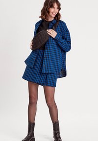 Chemise surdimensionnée à carreaux bleus et noirs, associée à une mini-jupe. Sac à taille matelassé noir, chemise noire en dessous et bottines brunes.