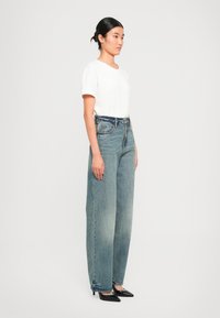 Donna in piedi di lato che indossa una maglietta bianca a maniche corte, jeans larghi a vita alta blu e décolleté neri a punta su uno sfondo neutro.