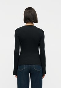 Γυναίκα με ίσια σκούρα μαλλιά που φοράει εφαρμοστή μαύρη μπλούζα με μακριά μανίκια και σκούρα μπλε τζιν, στέκεται με την πλάτη προς την κάμερα.