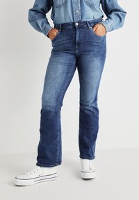 Jeans de mezclilla azul con diseño de cintura alta, pierna recta y bolsillos delanteros, combinados con zapatillas blancas para un look casual.