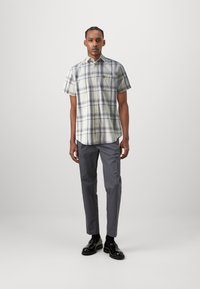 Camisa de plaid de manga corta en tonos claros con líneas oscuras, con un bolsillo en el pecho y un pequeño logo, combinada con pantalones grises y zapatos negros.